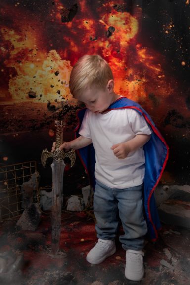A little boy with pale skin and blonde hair dressed as a superhero, holding a sword, with a fiery background.