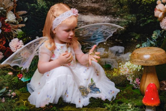 A little girl with pale skin and blonde hair dressed as a fairy, against a magical forest background.