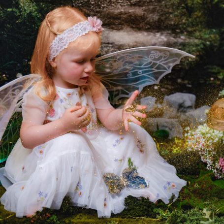A little girl with pale skin and blonde hair dressed as a fairy, against a magical forest background.