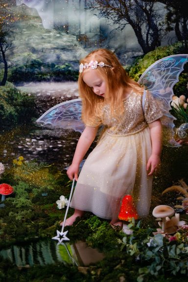 A little girl with pale skin and blonde hair dressed as a fairy, holding a magic wand, against a magical forest background.