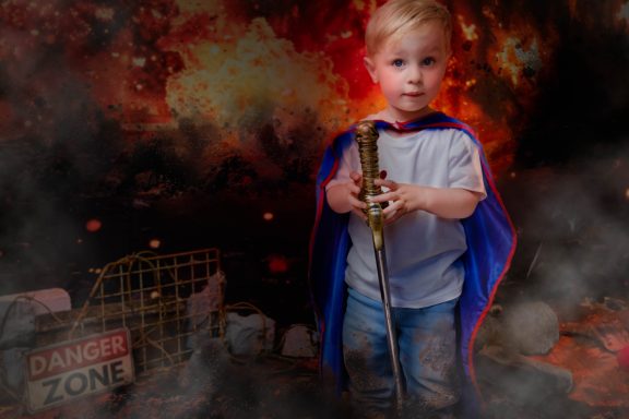 A little boy with pale skin and blonde hair dressed as a superhero, holding a sword, with a fiery background.