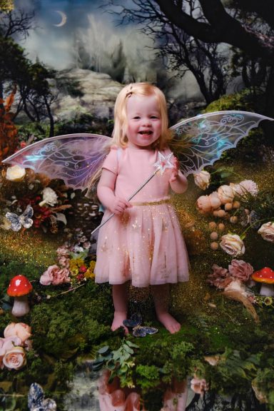 A little girl with pale skin and blonde hair dressed as a fairy, holding a magic wand, against a magical forest background.
