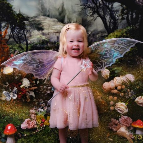 A little girl with pale skin and blonde hair dressed as a fairy, holding a magic wand, against a magical forest background.