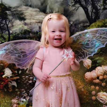 A little girl with pale skin and blonde hair dressed as a fairy, holding a magic wand, against a magical forest background.