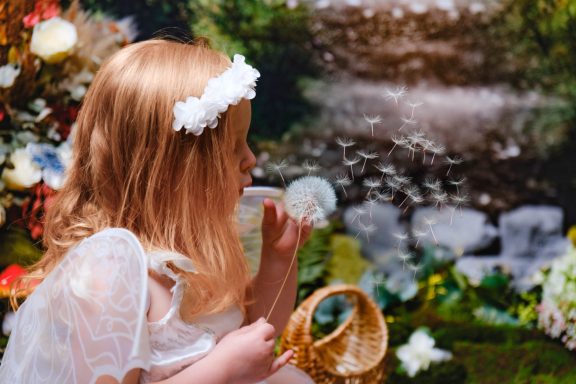 A little girl with pale skin and blonde hair dressed as a fairy, holding and blowing a dandelion clock, against a magical forest background.