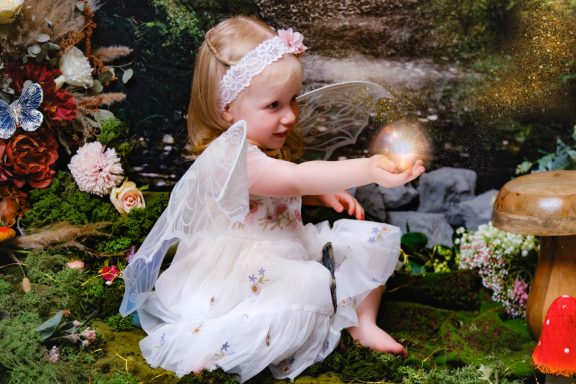 A little girl with pale skin and blonde hair dressed as a fairy, holding an orb of light, against a magical forest background.