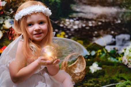 A little girl with pale skin and blonde hair dressed as a fairy, holding an orb of light, against a magical forest background.