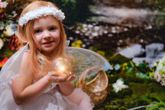 A little girl with pale skin and blonde hair dressed as a fairy, holding an orb of light, against a magical forest background.