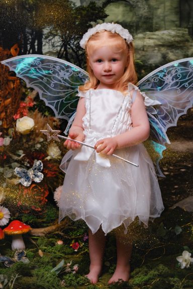 A little girl with pale skin and blonde hair dressed as a fairy, holding a magic wand, against a magical forest background.