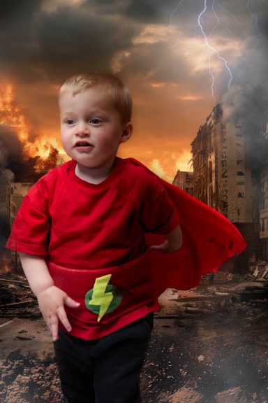 A little boy with pale skin and blonde hair dressed as a superhero, with a background of a destroyed city and lightening.