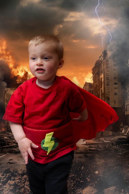 A little boy with pale skin and blonde hair dressed as a superhero, with a background of a destroyed city and lightening.