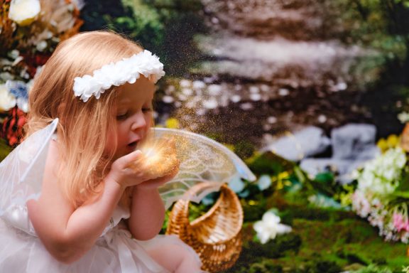 A little girl with pale skin and blonde hair dressed as a fairy, holding an orb of light, against a magical forest background.