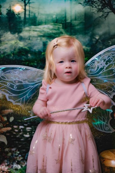 A little girl with pale skin and blonde hair dressed as a fairy, holding a magic wand, against a magical forest background.