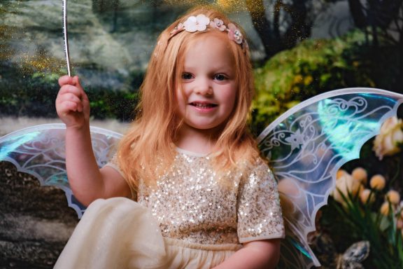 A little girl with pale skin and blonde hair dressed as a fairy, holding a magic wand, against a magical forest background.