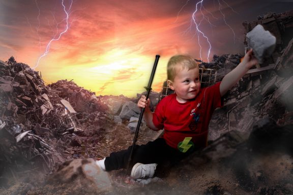 A little boy with pale skin and blonde hair dressed as a superhero, holding a sword, with a background of a destroyed city and lightening.