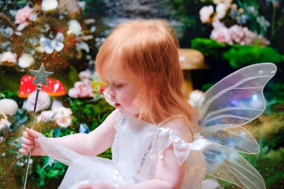 A little girl with pale skin and blonde hair dressed as a fairy, holding a magic wand, against a magical forest background.
