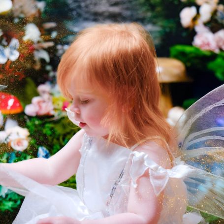 A little girl with pale skin and blonde hair dressed as a fairy, against a magical forest background.