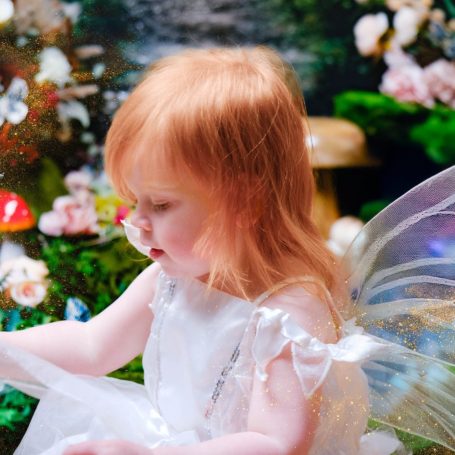 A little girl with pale skin and blonde hair dressed as a fairy, against a magical forest background.