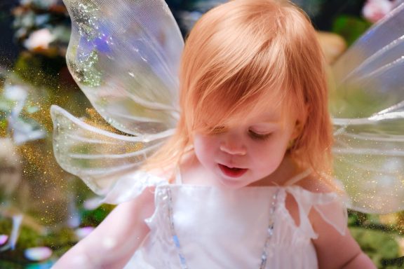 A little girl with pale skin and blonde hair dressed as a fairy, against a magical forest background.