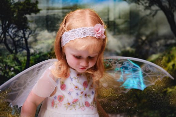A little girl with pale skin and blonde hair dressed as a fairy, against a magical forest background.
