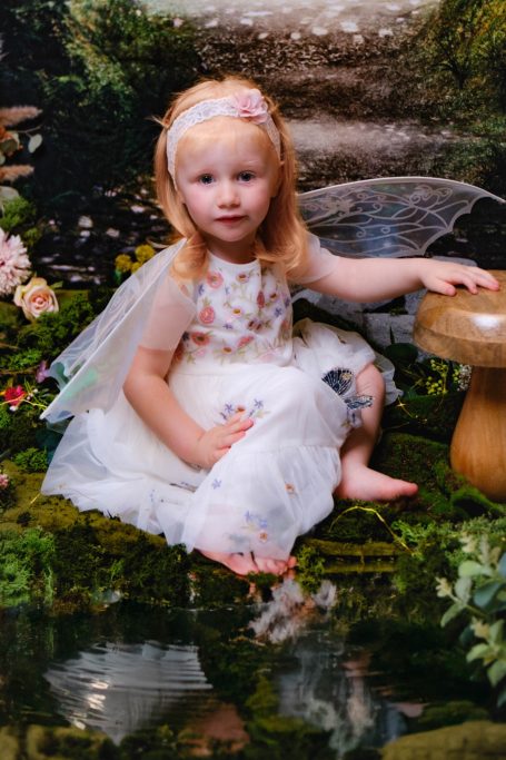 A little girl with pale skin and blonde hair dressed as a fairy, against a magical forest background.