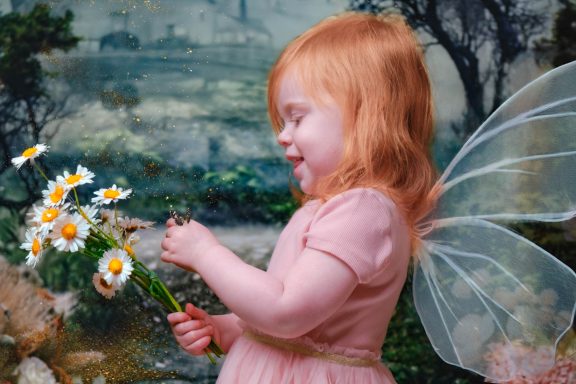 A little girl with pale skin and blonde hair dressed as a fairy, holding a bunch of daisies, against a magical forest background.