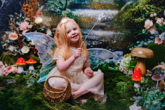 A little girl with pale skin and blonde hair dressed as a fairy, holding a magic wand, against a magical forest background.