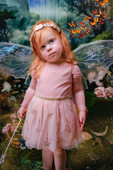 A little girl with pale skin and blonde hair dressed as a fairy, holding a magic wand, against a magical forest background.